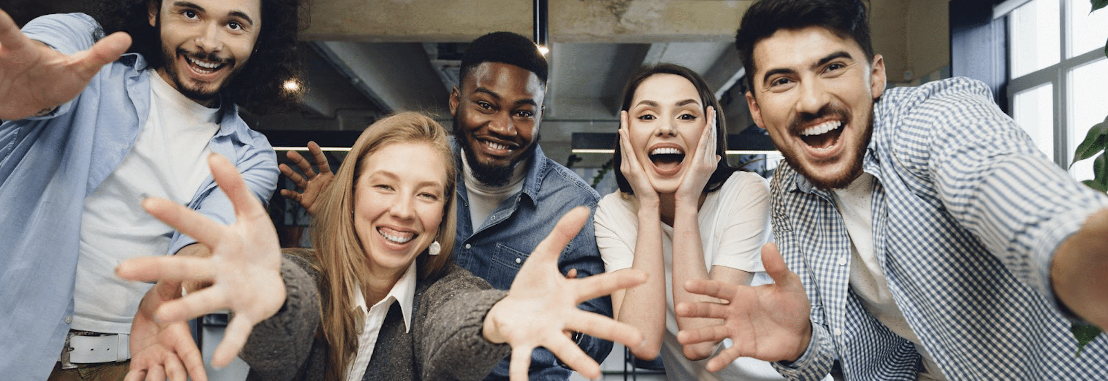 Groupe diversifié de cinq jeunes professionnels riant et tendant les mains vers la caméra dans un bureau lumineux