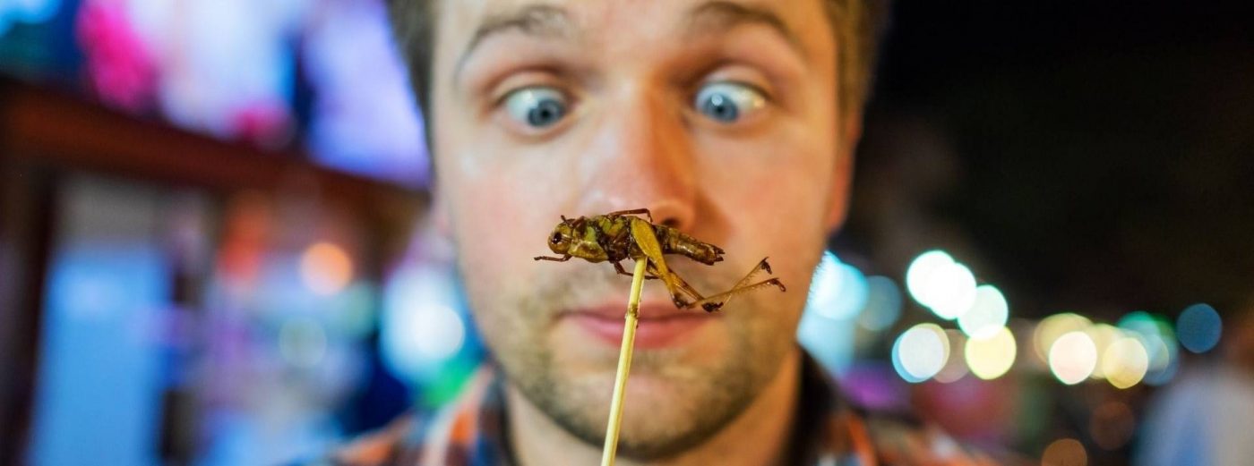 Homme exprimant la surprise face à un insecte grillé sur un bâton, prêt à être dégusté